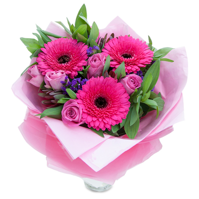 I picture this bouquet as a playful yet graceful composition made for intimate celebrations. Three fuchsia Gerbera daisies dominate the view, their bold, circular blooms layered with numerous petals and centred by dark discs that anchor the palette. Nestled close are several roses in a muted lavender-pink or dusty rose, each bud tightly coiled and soft to the touch, offering a tender contrast to the Gerberas' wide faces. Interspersed are tiny sprays of purple statice that lend an airy, meadow-like texture and a cool punctuation to the warm pinks. The greenery-salal or eucalyptus-like leaves-sits robustly behind the flowers, glossy and shaped to provide volume and a refreshing scent that evokes early-morning trims at the shop. The bouquet's wrapping is deliberate and elegant: an inner cushion of matte pale pink tissue envelops the stems, while an outer veil of sheer fuchsia-pink cellophane, slightly crinkled, catches light and gives the presentation a subtle sparkle. In the lower frame a clear glass vase rim is visible, suggesting the arrangement is ready to be set down on a hallway table or beside a favourite armchair. The overall mood is warm and reassuring, suitable for a thoughtful anniversary gift, a Mother's Day surprise, or a cheerful bedside pick-me-up. Working regularly across Slough-delivering around Slough High Street and near Herschel Park-I place these bouquets where their bright, optimistic colours will make the most immediate emotional impact.