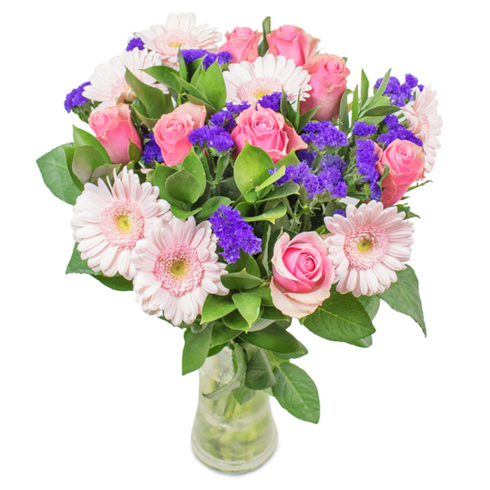 Nestled within a clear glass vase and set against a crisp white background, this exquisite floral bouquet showcases the skill of Slough's local florists. At its core are roughly seven to nine medium-pink roses, some with petals still gently curled and others just beginning to blossom, their iconic spirals evoking cherished memories along the Jubilee River. Surrounding these are five to seven pale pink gerbera daisies with broad, radiating petals and subtle cream centres, reminiscent of fresh blooms spotted in gardens off Stoke Poges Lane. Vivid purple statice or limonium punctuates the bouquet, introducing a lively pop of colour and textural contrast that brightens the ensemble like the seasonal displays at Wexham Park. Deep green, glossy foliage forms the base, suggesting lushness and vitality, evoking the peaceful shade of Langley Park woodlands. The stems and crystal-clear water in the transparent vase hint at freshness and the promise of a beautiful day. This arrangement-balancing classic and contemporary style-makes an ideal gift for birthdays, anniversaries, Mother's Day, or for adorning homes and workspaces across Slough. Every element is chosen not only for beauty but also to capture the spirit of cherished local moments.